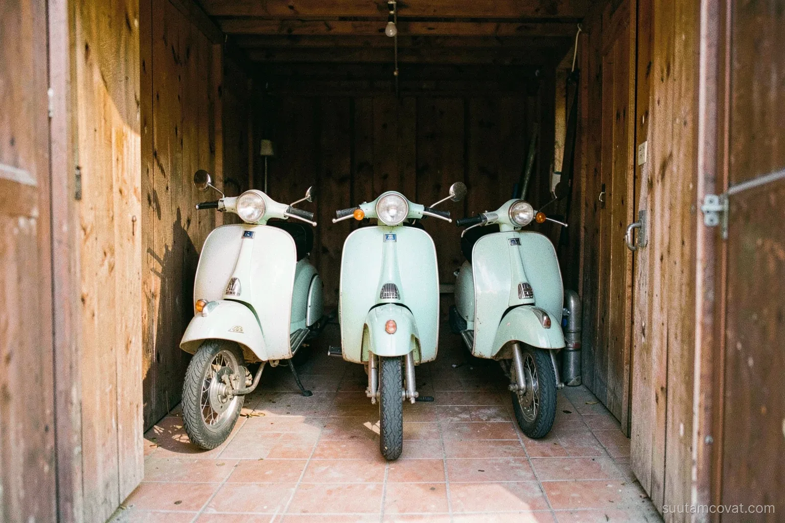 Three vintage Italian scooters in pastel colors lined up inside a narrow wooden garage, viewed from the open doorway, morning light falling across tiled floor, film grain