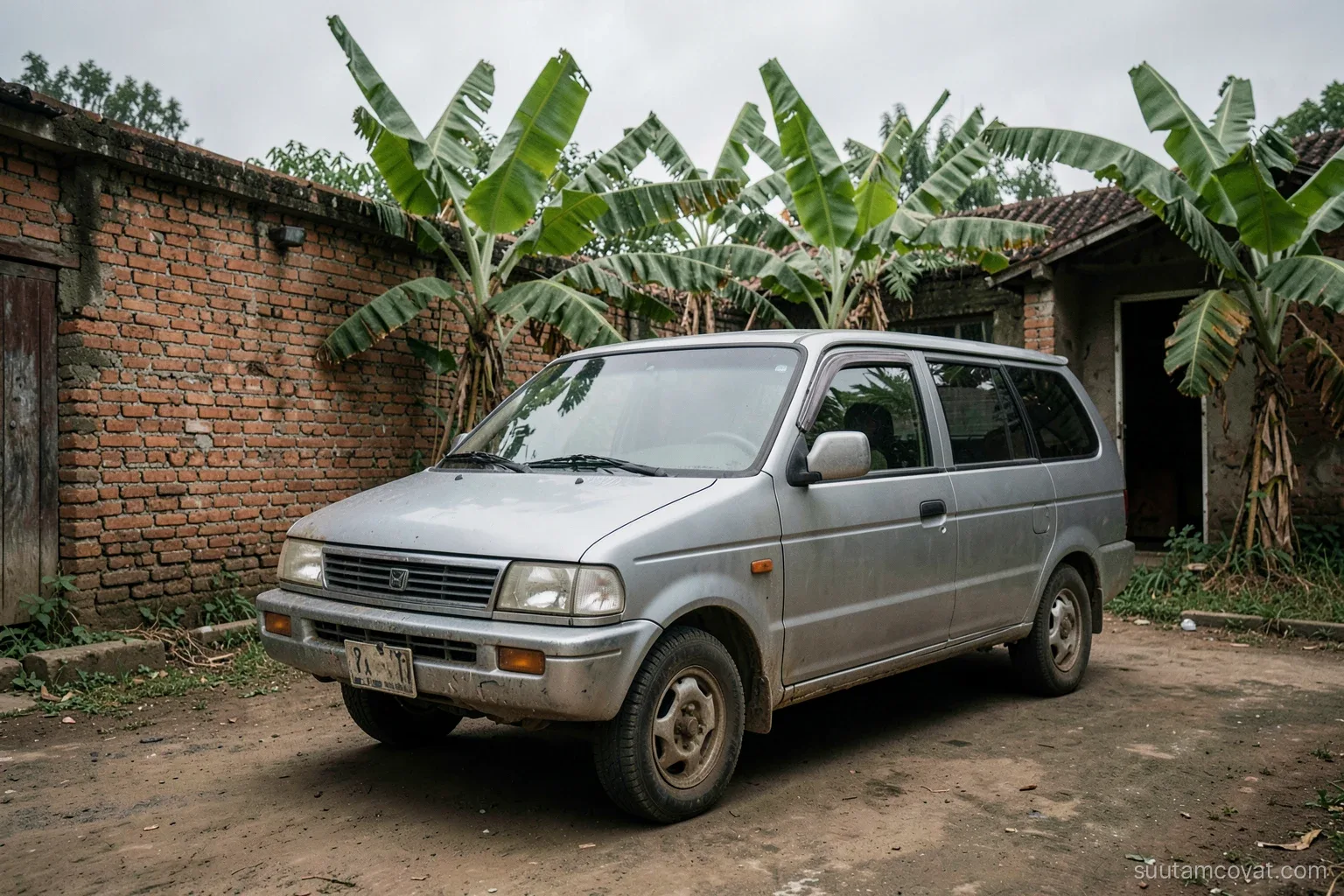 A boxy silver minivan from the late nineties parked in a rural Vietnamese courtyard beside a brick wall and banana trees, wide shot, overcast soft light, faded colors