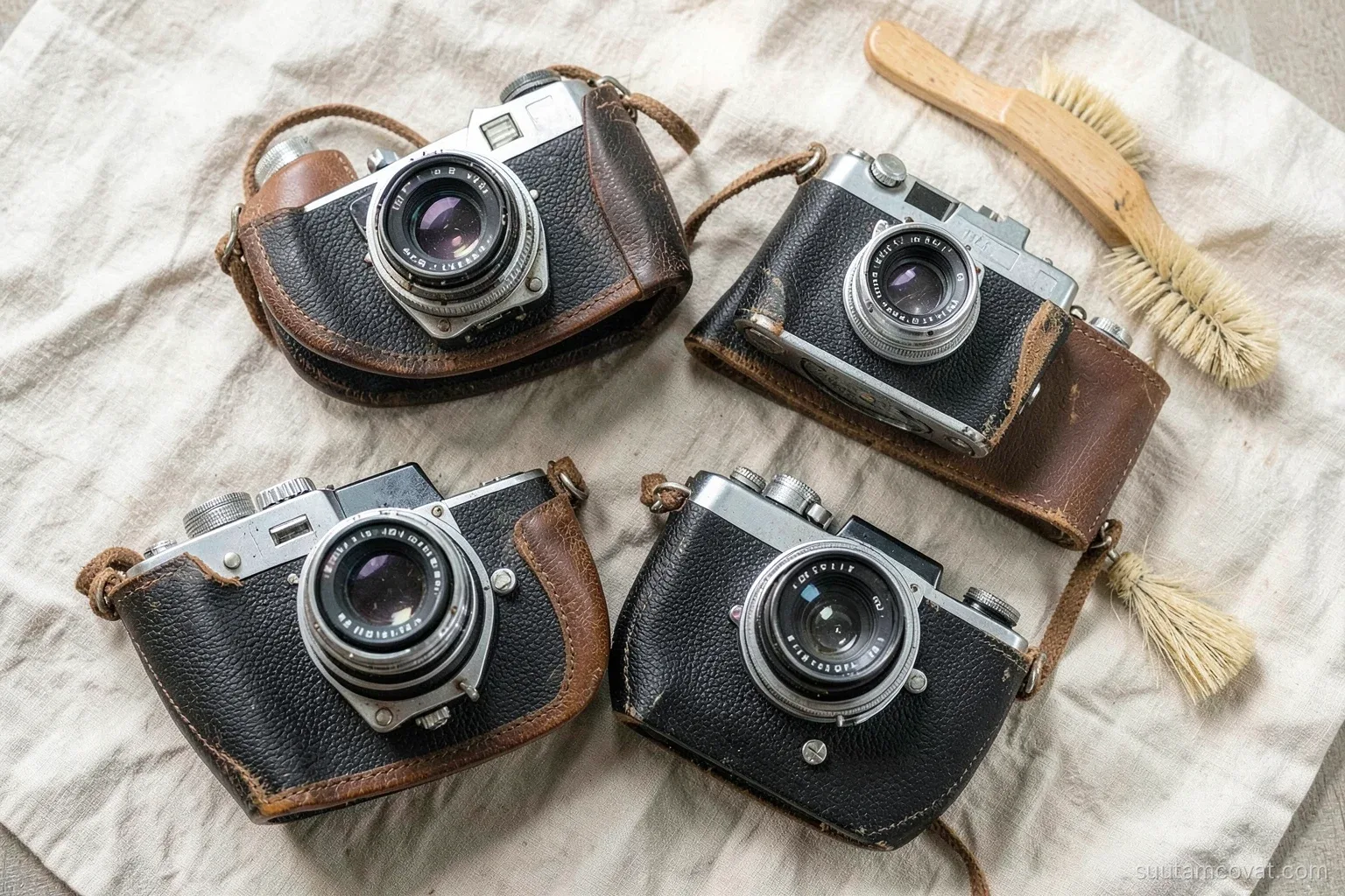 Top-down view of several old film cameras with leather covers and chrome lenses arranged on a linen cloth beside a soft brush and a blower, gentle window light